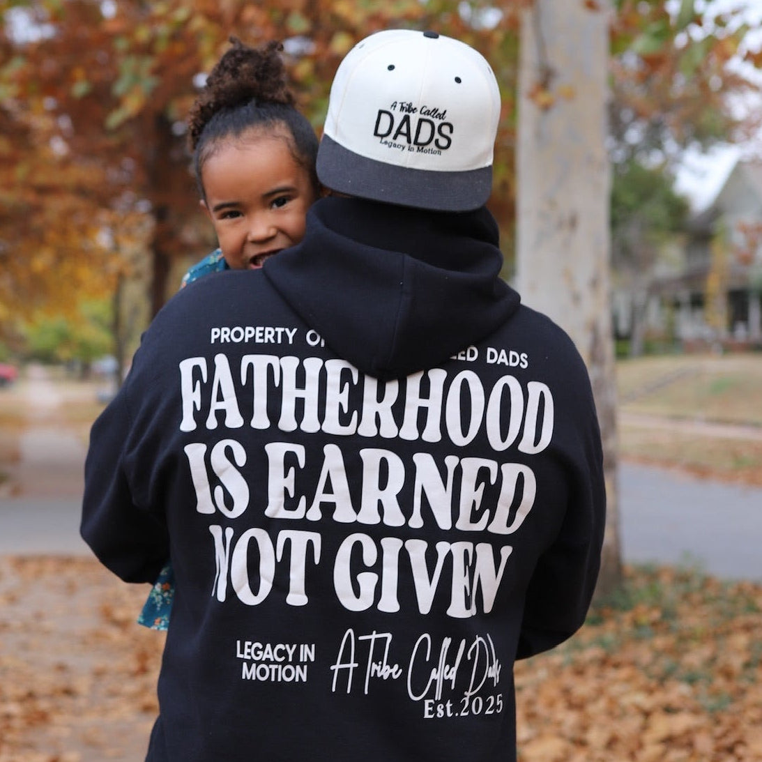 Person wearing a hoodie with 'Fatherhood is Earned Not Given' text, standing outdoors with trees in the background.